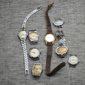 May include: A collection of vintage wristwatches with various designs and bands. The watches feature silver and gold tones, with leather and metal bands. The watch faces display different styles, including round, rectangular, and oval shapes. The watches are arranged on a gray fabric surface.