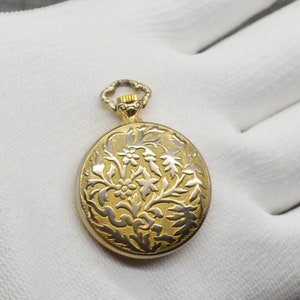 Peut inclure: Une montre de poche dor&eacute;e avec un motif floral sur le cadran. La montre a un fond blanc avec un motif floral dor&eacute;.