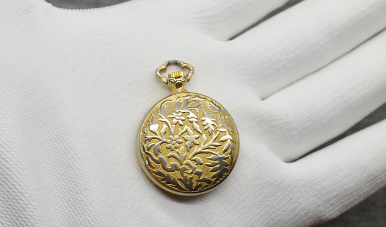 Peut inclure: Une montre de poche dor&eacute;e avec un motif floral sur le cadran. La montre a un fond blanc avec un motif floral dor&eacute;.