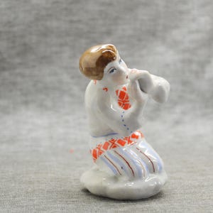 Könnte beinhalten: Eine Porzellanfigur einer Person, die ein Musikinstrument spielt. Die Figur ist in Weiß, Rot, Blau und Braun bemalt. Die Person trägt ein traditionelles Trachtenkleid mit einem rot-blau gemusterten Rock.