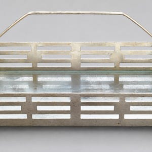 Puede incluir: Una cesta rectangular de metal plateado con un diseño de listones y recortes horizontales en los cuatro lados. La cesta incluye un asa articulada que se arquea sobre la parte superior y una base plana y reflectante en el interior.