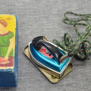 Op de afbeelding: Vintage elektrisch strijkijzer met een turquoise basis en zwarte handgreep, op een metalen bakje. Een blauwe doos met een vrouw in een groene jurk staat links. Een groen en geel snoer is opgerold.