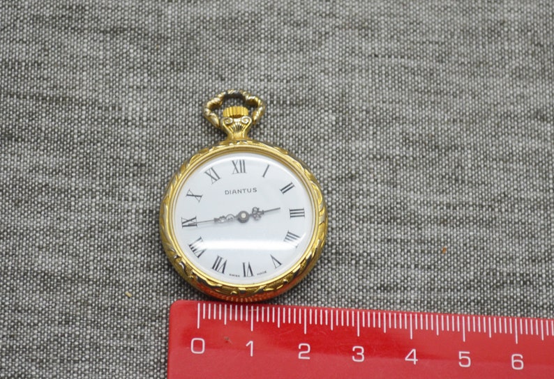 Peut inclure: Une montre de poche dor&eacute;e avec un cadran blanc et des chiffres romains noirs. Le cadran de la montre indique "DIANTUS" et "Swiss Made". La montre mesure environ 5 cm de diam&egrave;tre.