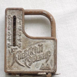 Puede incluir: Una báscula de resorte manual vintage en color marrón y plata con una ventana de medición vertical marcada de 0 a 5 kg. La cara metálica presenta texto cirílico en relieve "КИЕВСКИЙ СУВЕНИР" y un pequeño logotipo triangular.