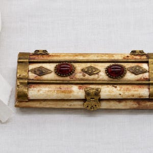 Puede incluir: Una caja rectangular con una superficie texturizada de color crema y patrones moteados marrones. Está decorada con adornos de metal de latón, dos piedras cabujón rojas ovaladas y tres pequeñas placas de latón en forma de diamante.