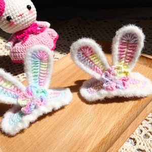May include: Two crocheted rainbow bunny ear hair clips with white fur trim and a pearl bead in the centre. The hair clips are on a wooden surface.