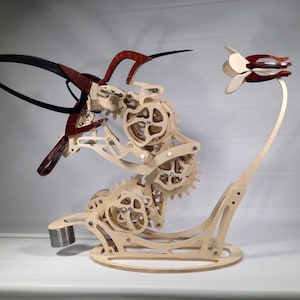 May include: A wooden kinetic sculpture of a hummingbird with intricate gears and a flower. The sculpture is made of natural wood with red accents and is mounted on a wooden base.