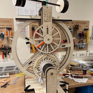 Puede incluir: Un reloj de madera con engranajes, ruedas y un peso en forma de barra en la parte superior. El reloj tiene el texto "ICTP SCI FAB LAB" y "The Leonardo Da Vinci Clock by FAB LAB". El reloj está sobre un banco de trabajo con herramientas.
