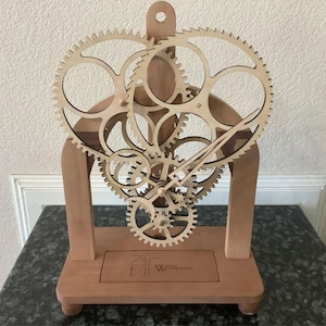 Puede incluir: Un reloj de madera con un diseño en forma de corazón y engranajes intrincados. El reloj está hecho de madera marrón claro y tiene una pequeña placa en la base con el texto "Time Works Wonders".