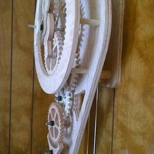 May include: A wooden clock mechanism with visible gears and a pendulum. The clock is made of light-colored wood and is mounted on a wooden wall. The gears are intricately designed and interlock to drive the clock's movement.