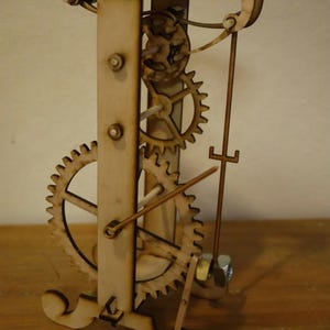 Può includere: Un modello in legno di un meccanismo di orologeria con ingranaggi e un pendolo. Il modello è in legno marrone chiaro e ha un design semplice.