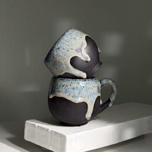 May include: Two handmade ceramic mugs stacked on a white rectangular platform. The mugs are dark brown with a blue and white glaze dripping down the sides. One mug is upside down on top of the other, showcasing the unique design.