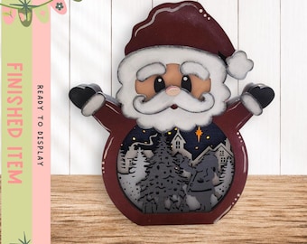 May include: A layered wooden Santa Claus decoration featuring a deep red hat and coat with a white beard. The center cutout displays a dark blue winter night scene with a Christmas tree, houses, and a bright yellow star, creating a dimensional effect.