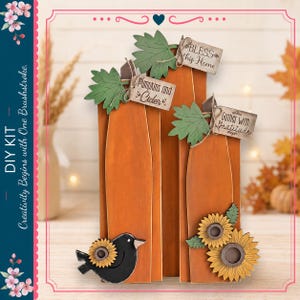 May include: A DIY kit featuring three tall orange wooden pumpkin cutouts. Each pumpkin is adorned with a green leaf and a small rectangular tag reading Bless This Home, Pumpkins and Cider, or Gather with Gratitude. Includes a black bird and sunflowers.