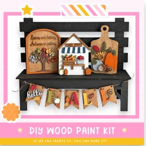 May include: A DIY wood paint kit featuring wooden fall-themed decorations. Includes a tree, a farm stand, a cutting board with pumpkins, and a banner that says "hello FALL". The kit is displayed on a black bench.