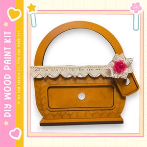 May include: A wooden DIY paint kit in the shape of a basket. The basket is light brown with a handle and a decorative lace trim. A pink flower with a gem is attached to the lace. The text "DIY WOOD PAINT KIT" is visible.