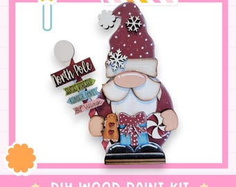 May include: A wooden gnome figurine DIY paint kit featuring a maroon hat with white polka dots and snowflakes, a white beard, and a signpost labeled North Pole, Santa's Workshop, Reindeer Games, and Elf Village. It holds a gingerbread man and gift.