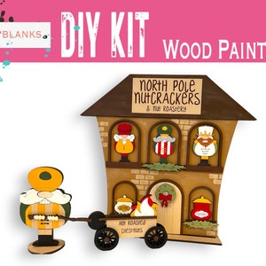 May include: DIY wood paint kit featuring a North Pole Nutcrackers & Nut Roastery building with a nutcracker figurine pulling a cart. The building has three windows with nutcracker figures inside and a sign that reads "North Pole Nutcrackers & Nut Roastery". The cart has a sign that reads "Hot Roasted Chestnuts".