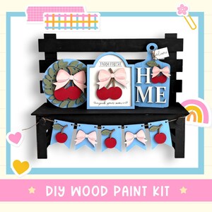 May include: A DIY wood paint kit featuring a black bench with decorative wooden pieces. The pieces include a wreath, a sign that says "Farm Fresh", a cutting board that says "Home", and a banner, all with cherry and bow accents.