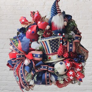 May include: A patriotic wreath decorated with red, white, and blue ribbons, stars, and a gnome wearing a red, white, and blue hat. The wreath is decorated with a top hat with a red, white, and blue design.