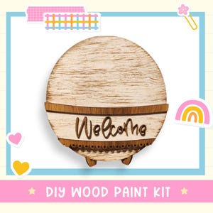 May include: A round wooden DIY paint kit with the word "Welcome" in a script font. The kit has a light wood grain finish with a dark brown border and decorative trim. The text "DIY WOOD PAINT KIT" is at the bottom.