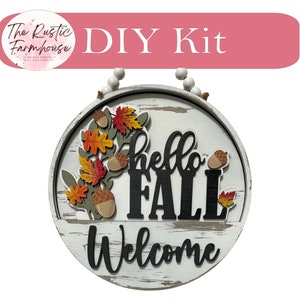 May include: DIY kit for a round wooden sign with the words "hello fall" and "welcome" in black lettering. The sign is decorated with fall leaves and acorns.