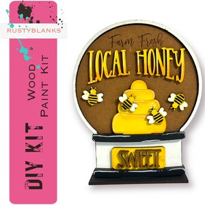 May include: DIY wood paint kit featuring a round wooden sign with the text "Farm Fresh Local Honey" and a beehive with bees. The sign has a base with the word "Sweet" on it.
