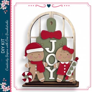 May include: A decorative wooden DIY kit featuring a green cutting board shape with the word JOY in white letters. It includes two gingerbread figures, one wearing a red Santa hat and scarf, the other a red dress and bow, plus a candy cane and rolling pin.
