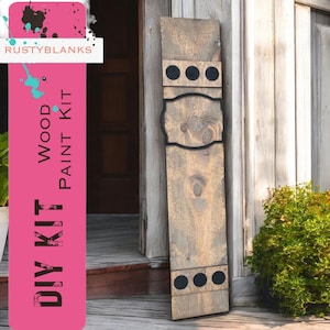 May include: DIY wood paint kit with a rustic wooden sign featuring a black metal frame and three black circles. The sign is leaning against a wooden door.