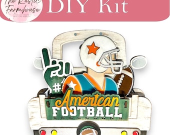 May include: A wooden DIY kit featuring a white truck cutout with a football player wearing a helmet, a green foam finger, and a brown football. The truck bed displays the text American FOOTBALL in yellow and white letters with yard line markings.