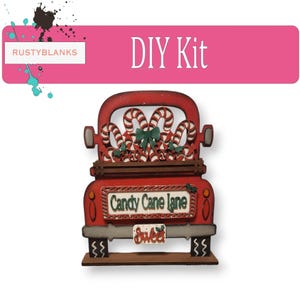 May include: A red wooden truck DIY kit featuring a truck bed filled with candy canes, a green bow, and a sign reading Candy Cane Lane. The truck has a license plate that says Sweet, a brown base, and a pink header with the text DIY Kit and RUSTYBLANKS.