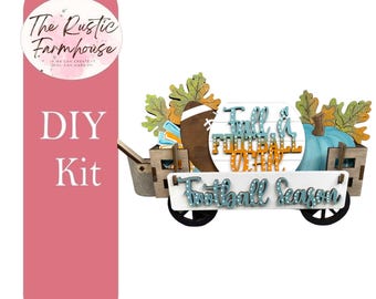 May include: A DIY kit featuring a wooden wagon filled with fall-themed decor, including a brown football, a light blue pumpkin, and green leaves. A white sign on the front reads Football Season, and a circular sign reads Fall & Football Dear.