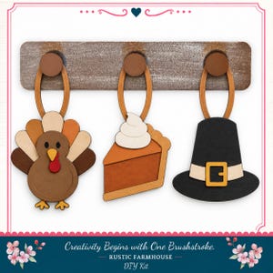 May include: A wooden wall rack with three pegs holding hanging ornaments: a brown turkey with a red wattle, a slice of pumpkin pie with white whipped cream, and a black pilgrim hat with a yellow buckle. Text reads: Creativity Begins with One Brushstroke.