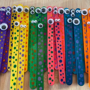 May include: A collection of colourful wooden craft sticks decorated with polka dots and googly eyes. The sticks are painted in various colours, including purple, yellow, green, red, blue, and orange.