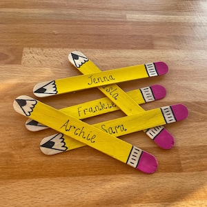 May include: Five yellow wooden craft sticks painted to look like pencils with pink eraser ends. Each stick has a name written on it: Jenna, Isla, Frankie, Archie, and Sara.