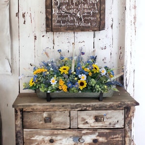 May include: A rustic wooden dresser with two drawers and a bottom shelf. The dresser is decorated with a floral arrangement in a gray planter. The flowers are a mix of yellow, blue, and white.
