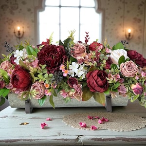 May include: A rectangular floral arrangement in a stone-colored trough. The arrangement features burgundy peonies, pink roses, white blossoms, and green foliage. The trough rests on a distressed, light blue wooden table.
