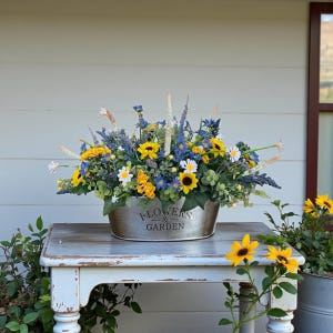 May include: A metal oval planter with the text FLOWERS & GARDEN embossed on the front, filled with an arrangement of artificial yellow sunflowers, white daisies, blue wildflowers, lavender sprigs, and green foliage.