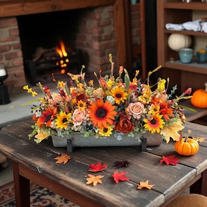 Puede incluir: Una mesa de madera rústica exhibe un vibrante arreglo floral otoñal en un recipiente metálico rectangular. El arreglo presenta girasoles, rosas y otras flores otoñales en tonos naranja, amarillo y rojo. Hojas de otoño decorativas y una pequeña calabaza completan la escena.