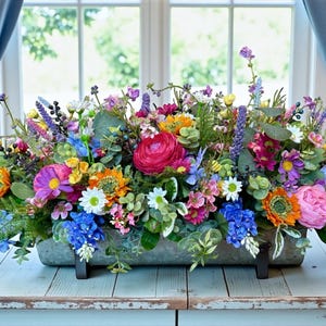 May include: A rectangular metal planter overflowing with a vibrant assortment of artificial flowers. The arrangement includes pink, purple, yellow, orange, and blue blooms, with green foliage. The planter sits on a light blue wooden surface.