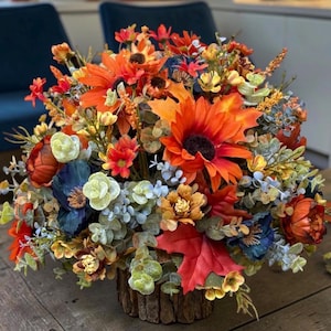 May include: A floral arrangement featuring orange sunflowers, deep red maple leaves, dark blue flowers, and light green eucalyptus foliage. The bouquet is contained within a cylindrical base with a textured, bark-like brown exterior finish.