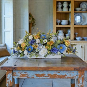 May include: A rectangular floral arrangement featuring a variety of flowers in shades of blue, yellow, and white. The flowers are arranged in a white wooden box, placed on a distressed wooden table with blue paint.