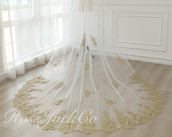 May include: A white tulle wedding veil with a gold embroidered floral pattern. The veil is long and flowing, with a scalloped edge.