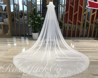 May include: A long, white bridal veil with a beaded edge. The veil is draped over a mannequin and is displayed on a tiled floor.