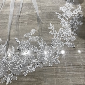 May include: White bridal veil with a floral design. The veil is made of tulle and has a delicate floral pattern embroidered on the edge.