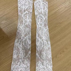May include: A pair of long white lace gloves featuring an intricate floral pattern throughout the fabric. The gloves have a delicate scalloped edge at the top and bottom openings, with a sheer, lightweight construction and a fine mesh base.