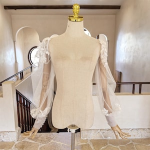 May include: A sheer, off-white bridal sleeve with floral lace detailing. The sleeves are long and feature a puffed design at the shoulder and wrist. The garment is displayed on a mannequin with a gold-colored top.