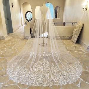 May include: A long, sheer white bridal veil with intricate floral lace detailing along the bottom edge. The veil is displayed on a mannequin, showcasing its length and delicate design. The text "Ross Jack Co" is visible on the veil.