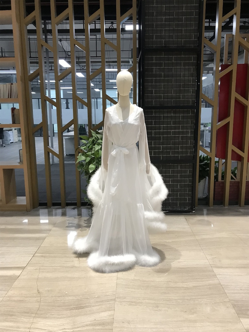 May include: A white sheer robe with a fluffy white trim around the hem and sleeves. The robe has a V-neck and a sash tie at the waist.