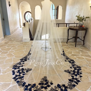 Op de afbeelding: Een witte tule bruids sluier met een marineblauwe en gouden bloemenkant rand. De sluier wordt getoond op een mannequin in een kamer met een stenen vloer.